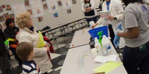 A woman and a child stand at a table with several people, including those in lab coats, in a room with tables and posters on the wall.